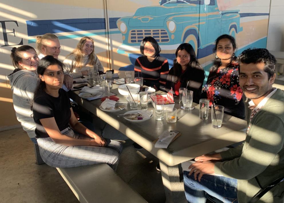 8 people sit around a picnic table with food and drinks in partial shade/sunlight