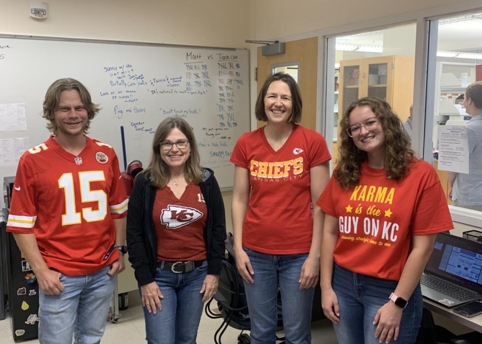 Four people stand in office wearing Chiefs shirts