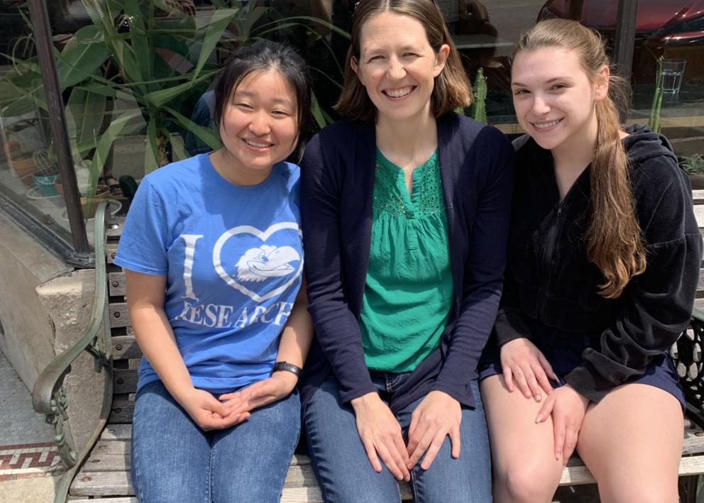 Three women sit on a bench outside smiling.