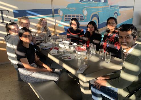 8 people sit around a picnic table with food and drinks in partial shade/sunlight