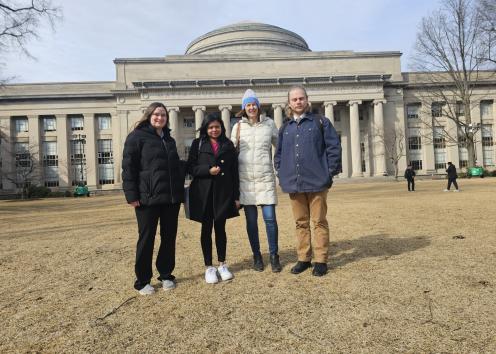 Four people in coats stand on lawn in front of MIT dome