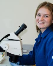 A woman smiles in front of a microscope wearing a lab coat